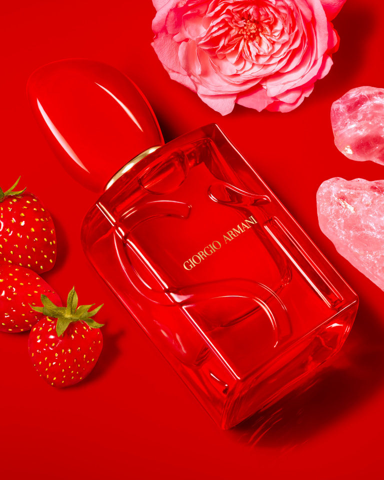 Photograph of a bottle of Giorgio Armani Sì resting on a red floor with a close-up of a slice of pear, vanilla pod and flower petals nearby