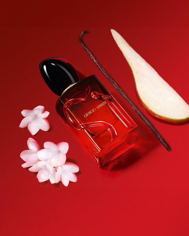 Photograph of a bottle of Giorgio Armani Sì resting on a red floor with a close-up of a slice of pear, vanilla pod and flower petals nearby